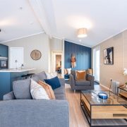 The open-plan living area is bright and inviting, with clean lines, generous seating and considered styling that elevates every detail. The statement kitchen, finished in rich blue cabinetry with a wood-grain feature wall, offers a beautifully appointed island — perfect for relaxed dining and effortless hosting. In the master suite, layered textures, a bold headboard and subtle geometric detailing create a serene retreat, complemented by a beautifully finished en-suite.