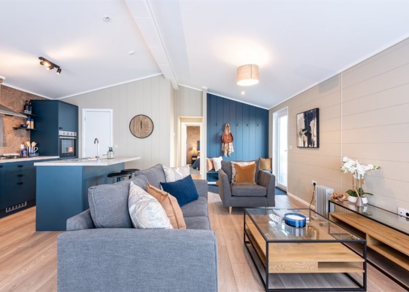The open-plan living area is bright and inviting, with clean lines, generous seating and considered styling that elevates every detail. The statement kitchen, finished in rich blue cabinetry with a wood-grain feature wall, offers a beautifully appointed island — perfect for relaxed dining and effortless hosting. In the master suite, layered textures, a bold headboard and subtle geometric detailing create a serene retreat, complemented by a beautifully finished en-suite.